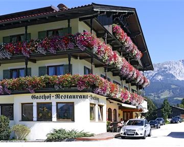 Ein traditionelles Gasthaus mit bunten Blumenstöcken. Im Hintergrund sind Berge und ein blauer Himmel zu sehen.