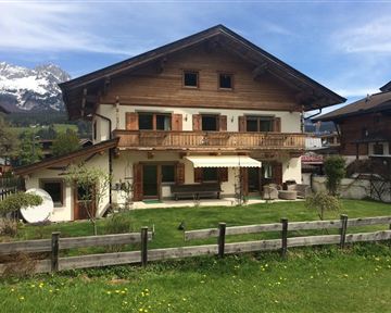 Ein schönes Holzhaus mit Balkon und Garten. Im Hintergrund sind die Berge zu sehen.