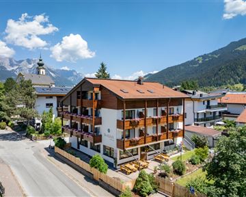 Ein charmantes Hotel mit Holzverkleidung und Balkonen, umgeben von grüner Natur. Im Hintergrund sind majestätische Berge und ein blauer Himmel zu sehen.