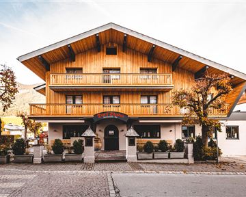 A traditional wooden house with several balconies and a spacious entrance area. The facade has a warm, inviting character and is surrounded by small trees.