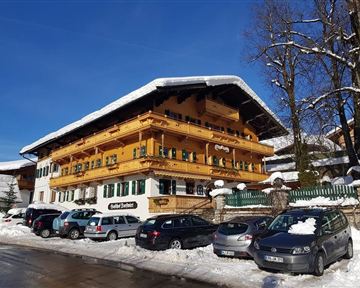 Ein charmantes, schneebedecktes Haus im alpinen Stil mit holzverzierten Balkonen. Umgeben von einer winterlichen Landschaft und geparkten Autos.