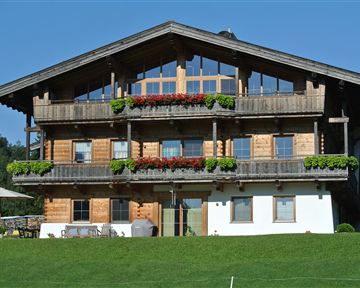 Ein traditionelles Holzhaus mit mehreren Balkonen und bunten Blumen. Es liegt in einer grünen Umgebung unter blauem Himmel.