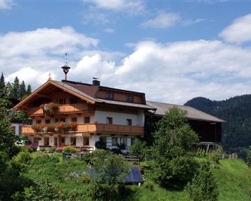Ein malerisches Haus im ländlichen Raum, umgeben von Bäumen und Wiesen. Im Hintergrund sind sanfte Berge und ein klarer Himmel zu sehen.