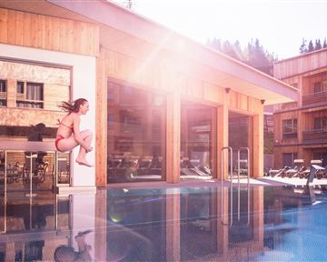 A girl jumps into a pool in front of a modern wooden building. The sun is shining, and it is a clear, beautiful day.