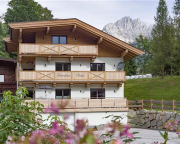 Ein schönes Holzhaus in den Bergen mit einem Balkon und umgeben von grüner Natur. Im Hintergrund sind beeindruckende Berge zu sehen.
