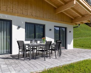 Eine moderne Terrasse mit einem Esstisch und Stühlen. Im Hintergrund ist ein grüner Hang und große Fenster sichtbar.