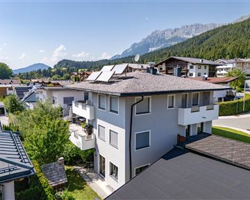 A modern house with several floors and solar panels. In the background, green mountains and a quiet residential area can be seen.