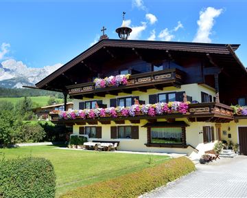 Ein charmantes alpenländisches Haus mit bunten Blumen auf dem Balkon. Im Hintergrund sind grüne Wiesen und hohe Berge zu sehen.