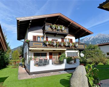Ein charmantes, zweistöckiges Haus mit Blumenbalkonen und einem schönen Garten. Im Hintergrund sind Berge und ein klarer Himmel zu sehen.