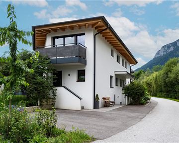 Ein modernes Haus mit Holzveranda und Garten, umgeben von Bäumen und Bergen. Die Zufahrtsstraße führt auf das Grundstück.