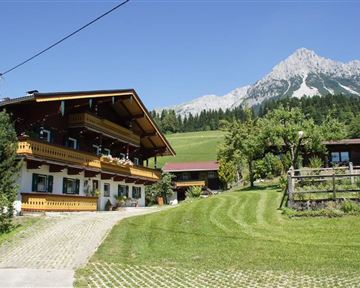 A traditional chalet with balconies in a picturesque mountain landscape. In the background, a majestic peak rises above green meadows and trees.