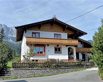 A cozy house in the mountains with wooden balustrades and a large garden. In the background, high mountains and a blue sky can be seen.