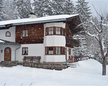 Ein charmantes Haus im Schnee, umgeben von schneebedeckten Bäumen. Die Szene vermittelt eine ruhige Winteratmosphäre.