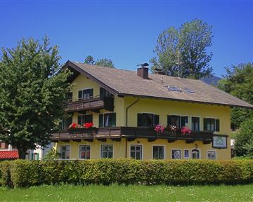 Ein sonniges Gebäude mit gelber Fassade und Balkonen, umgeben von Bäumen. An der Fassade sind Blumen Kästen mit bunten Pflanzen angebracht.