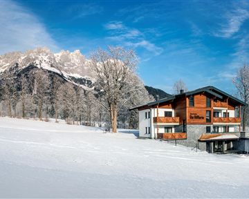 A charming house in a snow-covered landscape with majestic mountains in the background. The trees are frosty and the sky is clear and blue.