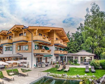 Ein schönes Hotel im alpenländischen Stil mit Holzverkleidung. Der Garten ist grün und einladend, mit Liegestühlen und einem Schwimmbecken.