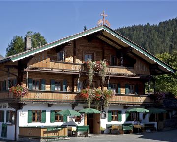 A traditional wooden house with colorful flowers and green awnings. In the background, forested hills can be seen.