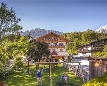 Ein malerisches Bergdorf mit traditionellen Holzhäusern und grünen Wiesen. Im Vordergrund gibt es einen Spielplatz und die beeindruckenden Berge im Hintergrund.
