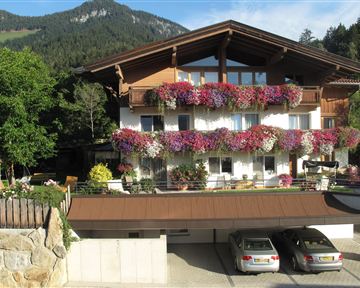 Ein malerisches Haus mit Blumenkästen und einer schönen Balkonansicht. Im Vordergrund sind zwei Autos vor einer Garage zu sehen, und im Hintergrund erstrecken sich Berge.