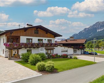 A charming country house with flower balconies, surrounded by green meadows and mountains. The sky is blue with a few clouds.
