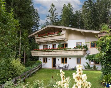Ein charmantes Holzhaus mit Balkon, umgeben von grünen Bäumen und bunten Blumen. Die Atmosphäre ist ruhig und einladend.