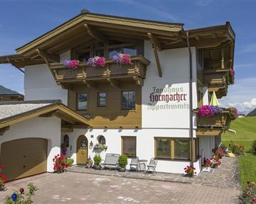 A mountain-style building with several balconies, adorned with colorful flowers. In the foreground, there are parking spaces and seating areas.