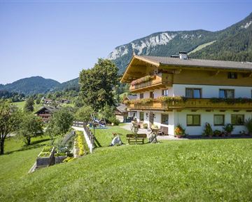 A picturesque house in a green landscape, surrounded by mountains. The sky is clear and the surroundings feel inviting and calm.