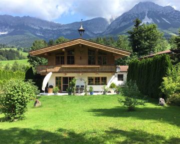 Ein schönes, traditionelles Holzhaus umgeben von grünem Rasen und Bäumen. Im Hintergrund erheben sich majestätische Berge unter einem klaren Himmel.