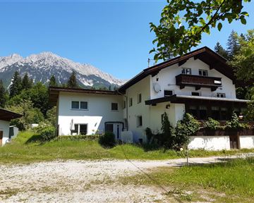 Ein schönes Gebäude inmitten grüner Wiesen und Bäume, mit majestätischen Bergen im Hintergrund. Der Himmel ist klar und blau.