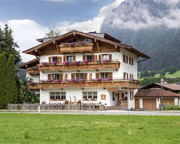 Ein charmantes Holzhaus mit Balkonen und bunten Blumen. Im Hintergrund sind Berge und ein bewölkter Himmel zu sehen.