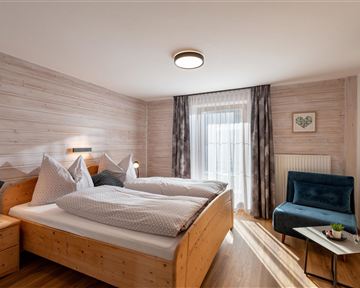 Ein gemütliches Schlafzimmer mit zwei Betten und holzernen Möbeln. Ein blauer Sessel und ein Fenster mit Vorhängen bieten einen einladenden Ausblick.
