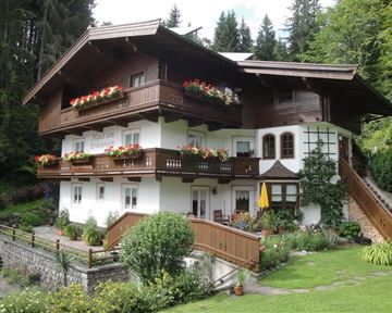Ein charmantes alpenländisches Haus mit Balkonen und blühenden Blumen. Umgeben von grüner Landschaft und Bäumen.