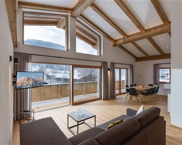 A modern living room with a wooden beam ceiling and large windows. There is a comfortable sofa and a table set up for sociable gatherings.