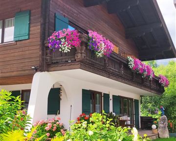 Ein traditionelles Holzhaus mit bunten Blumenbalkonen. Der Garten ist mit üppigen Pflanzen und Blumen gestaltet.