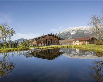 Ein malerisches Holzhaus am Rand eines klaren Teichs. Im Hintergrund sind beeindruckende Berge unter einem klaren Himmel zu sehen.
