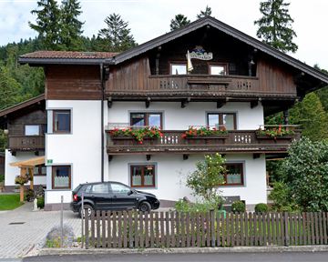 A two-story house in alpine style with a balcony and blooming windows. In the foreground, there is a car, and there is a small garden with a fence.