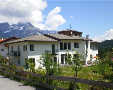 Ein modernes Haus mit Balkon, umgeben von grüner Landschaft und Blumen. Im Hintergrund sind die Berge sichtbar, unter einem blauen Himmel mit einigen Wolken.
