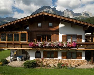 Ein traditionelles Holzhaus mit bunten Blumen auf dem Balkon. Im Hintergrund sind majestätische Berge und ein klarer Himmel zu sehen.