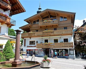 A charming building in alpine style with balconies and colorful flowers. In the foreground, there is a fountain and visitors are exploring the surroundings.