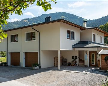 Ein modernes Haus in einer ländlichen Umgebung mit grünen Bergen im Hintergrund. Die Fassade ist hell und freundlich, umgeben von Bäumen.