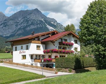 Ein malerisches Haus mit Blumenbalkonen, umgeben von grünen Wiesen. Im Hintergrund erheben sich beeindruckende Berge unter einem blauen Himmel.