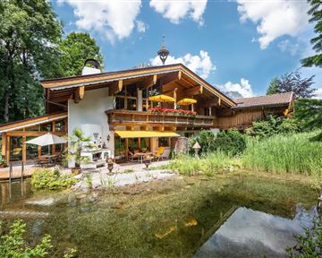 Ein charmantes Holzhaus mit einem schönen Garten und einem kleinen Teich. Im Hintergrund sind Bäume und ein strahlend blauer Himmel zu sehen.
