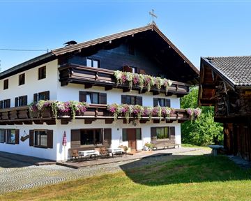 Ein malerisches Haus im alpenländischen Stil mit Balkonen, die mit bunten Blumen geschmückt sind. Der Himmel ist klar und die Umgebung ist grün und einladend.