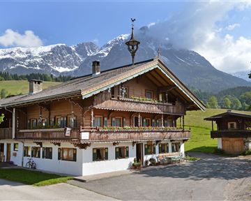 A traditional alpine house surrounded by green meadows and high mountains. Snow-capped peaks are visible in the background.