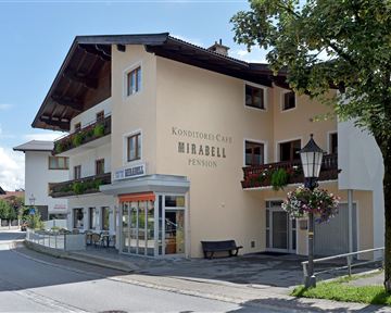A charming guesthouse decorated with balconies and flowers. The facade is bright and inviting, surrounded by trees and a peaceful street scene.