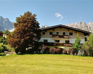 Ein malerisches Gebäude umgeben von einem grünen Garten und Bäumen. Im Hintergrund sind majestätische Berge zu sehen.