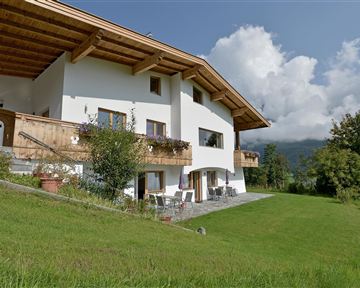 A charming house with a traditional wooden porch, surrounded by green meadows. In the background, gentle hills and a blue sky can be seen.