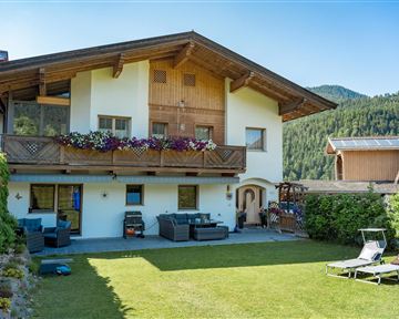 Ein gemütliches Haus mit einem Garten in den Bergen. Das Gebäude hat einen Balkon mit Blumen und ist von grünen Wiesen umgeben.