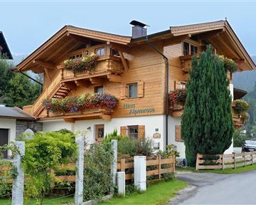 Ein charmantes, holzverkleidetes Haus mit Blumenbalkonen und gepflegtem Garten. Die Umgebung ist von grünen Hügeln und einem ruhigen Weg umgeben.