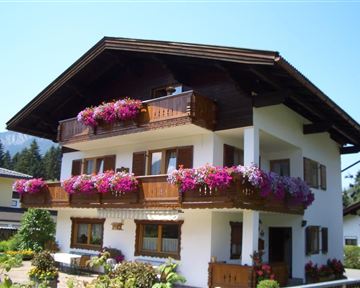 A charming house with blooming balconies and a bright blue sky. It is surrounded by green plants and trees.